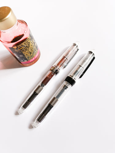Two Komamono Lab Fonte Transparent Fountain Pens with Ink Converters and a bottle of ink on a white surface.